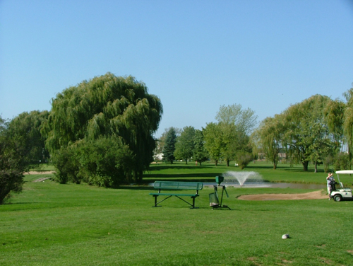 Course greens with a green bench in front of a pond with a fountain 